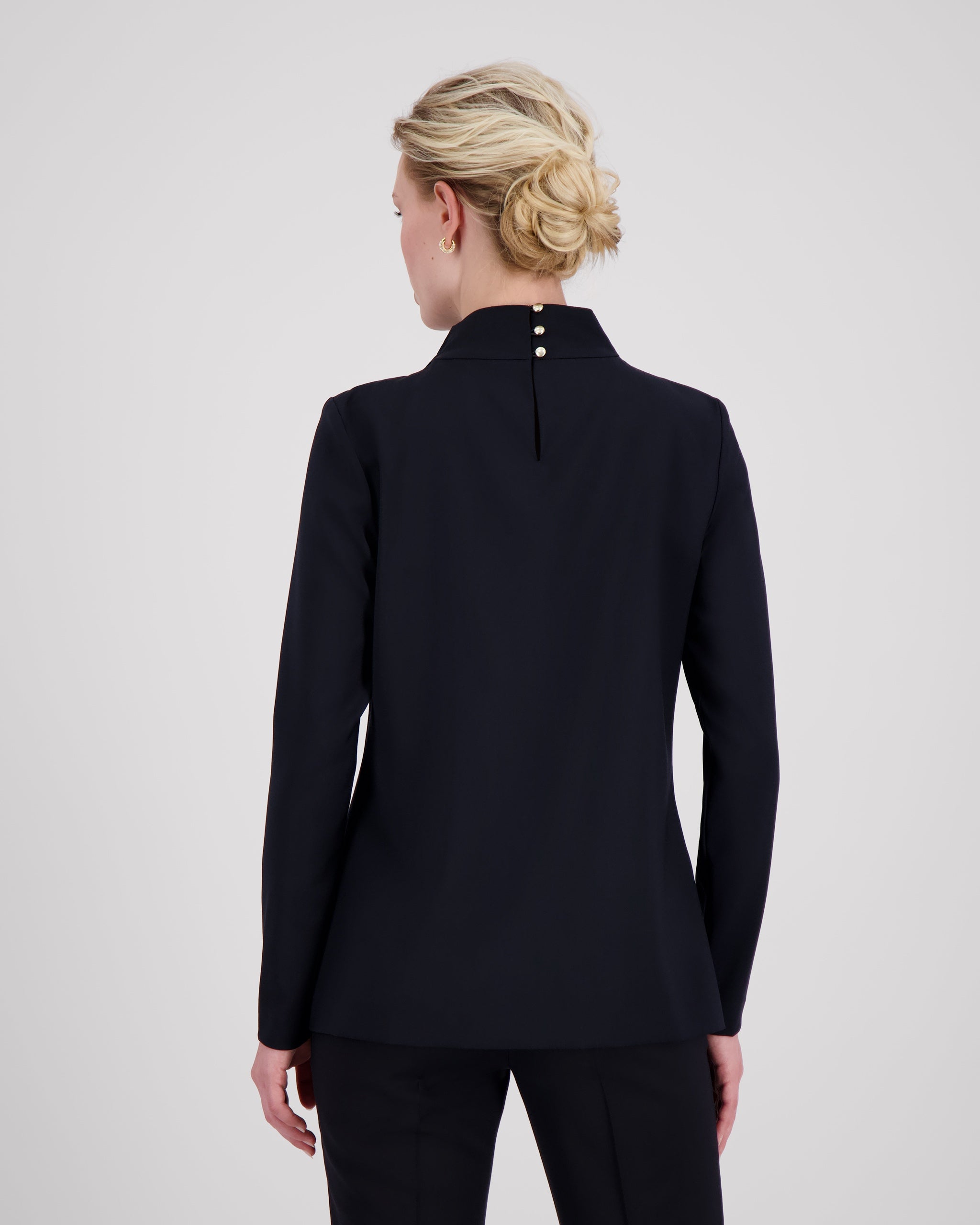 Top mit hochgeschlossenem, sanft drapiertem Stehkragen und feinem Rückenschlitz mit drei goldenen Knöpfen als dezentes, edles Detail. Zweifach umgeschlagene Ärmel und ein schmal abgesteppt Saum betonen die präzise Linienführung. Die körpernahe, weich fliessende Passform verbindet skulpturale Eleganz mit femininer Leichtigkeit.