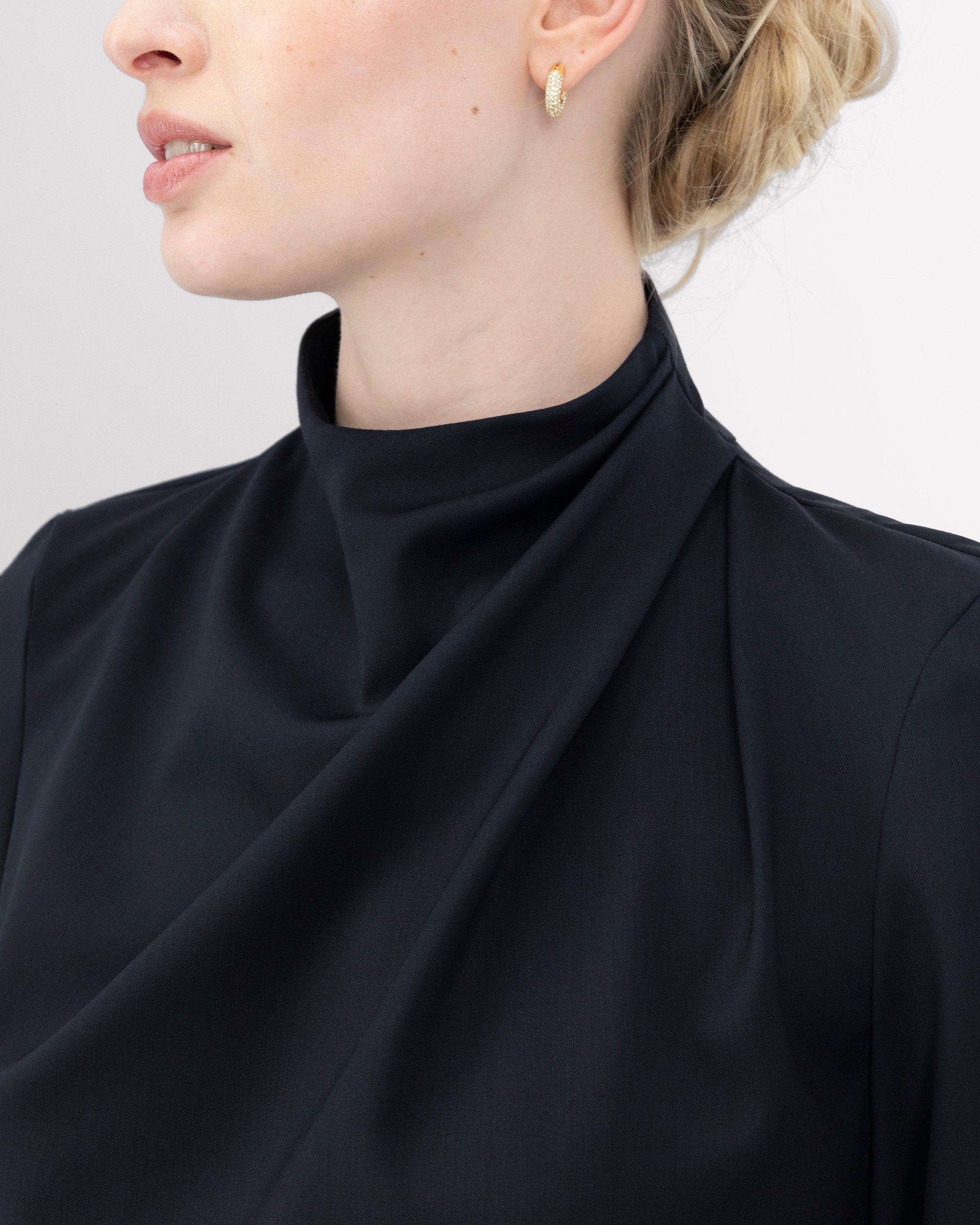 Top mit hochgeschlossenem, sanft drapiertem Stehkragen und feinem Rückenschlitz mit drei goldenen Knöpfen als dezentes, edles Detail. Zweifach umgeschlagene Ärmel und ein schmal abgesteppt Saum betonen die präzise Linienführung. Die körpernahe, weich fliessende Passform verbindet skulpturale Eleganz mit femininer Leichtigkeit.