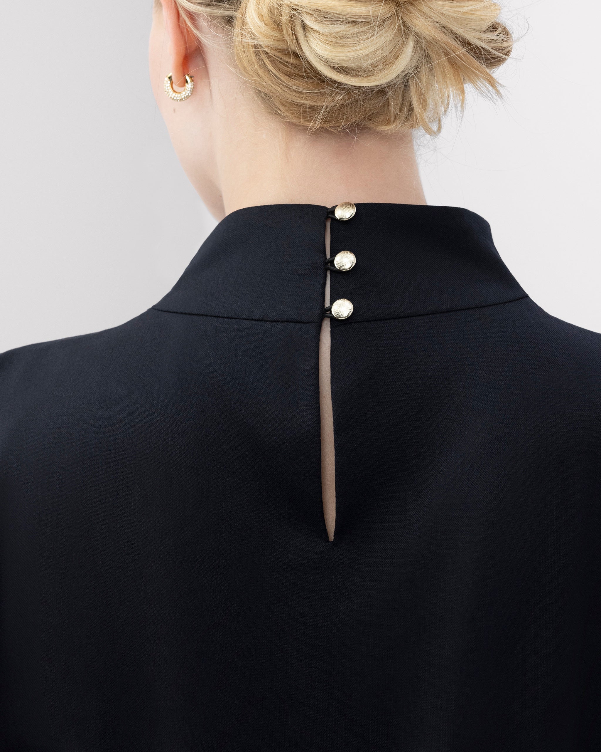 Top mit hochgeschlossenem, sanft drapiertem Stehkragen und feinem Rückenschlitz mit drei goldenen Knöpfen als dezentes, edles Detail. Zweifach umgeschlagene Ärmel und ein schmal abgesteppt Saum betonen die präzise Linienführung. Die körpernahe, weich fliessende Passform verbindet skulpturale Eleganz mit femininer Leichtigkeit.