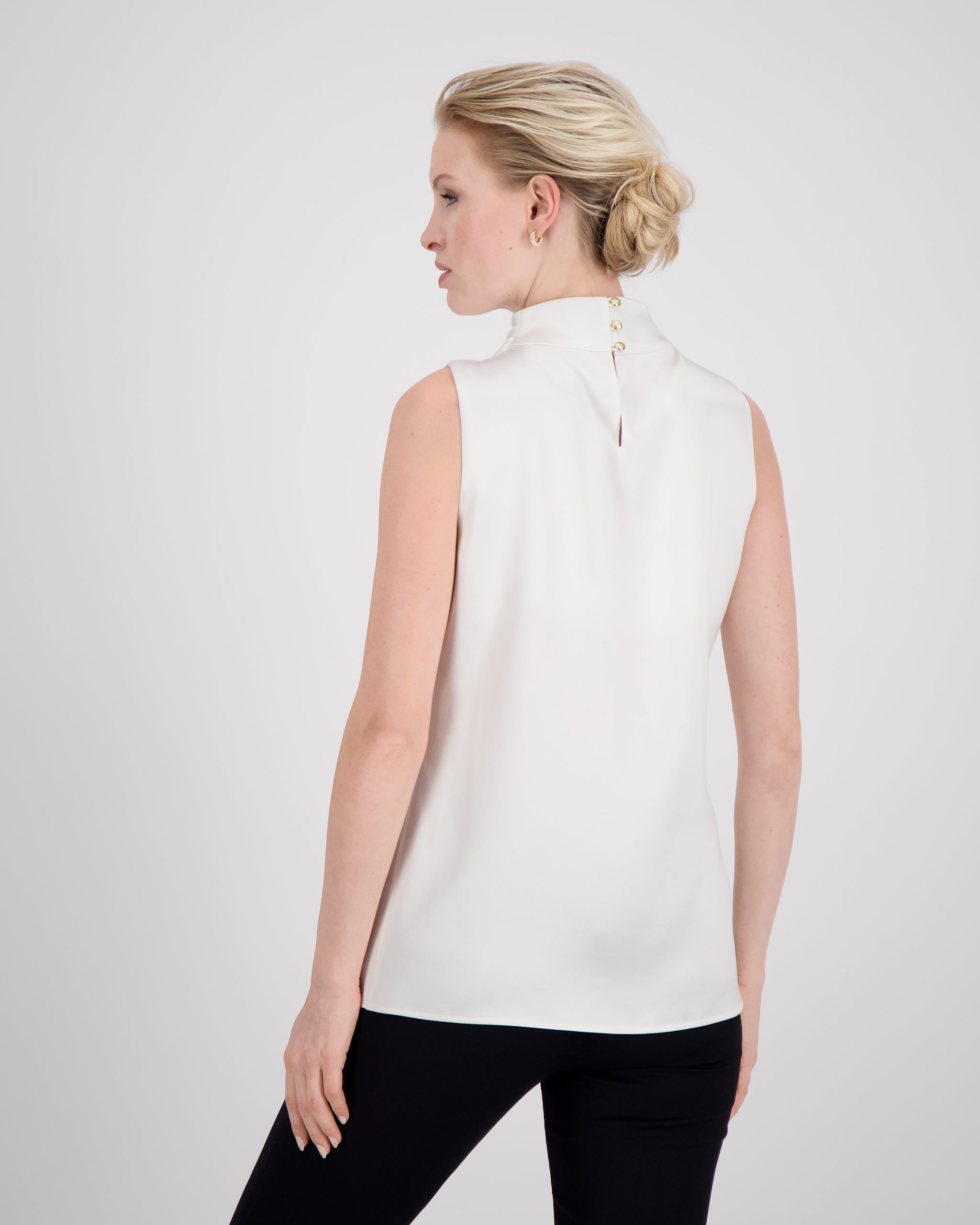 Asymmetrisch geschnittenes Top mit sanft drapierter Schulter für eine feminine, schwungvolle Silhouette. Der hochgeschlossene Stehkragen wird im Rückenteil durch einen feinen Schlitz und drei goldene Knöpfe mit filigranen Knopfschlingen akzentuiert. Ein edles Detail, das das puristische Design um eine subtile Note von Noblesse ergänzt.