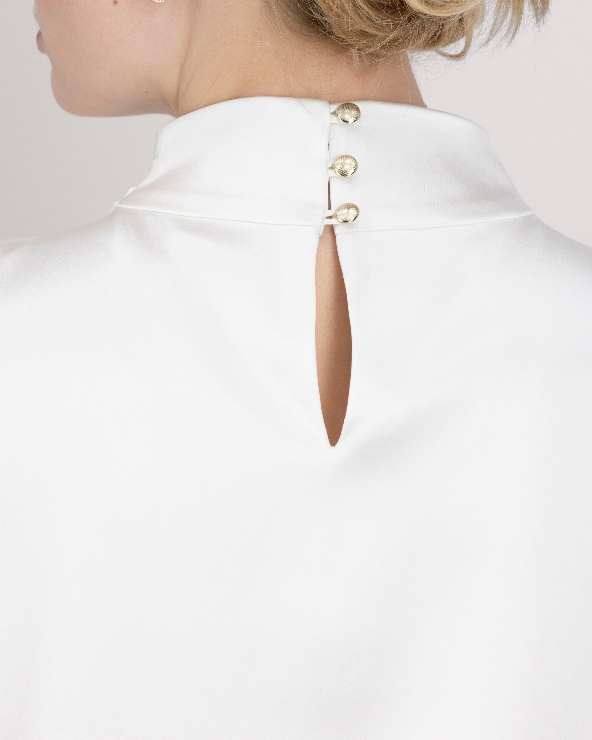 Asymmetrisch geschnittenes Top mit sanft drapierter Schulter für eine feminine, schwungvolle Silhouette. Der hochgeschlossene Stehkragen wird im Rückenteil durch einen feinen Schlitz und drei goldene Knöpfe mit filigranen Knopfschlingen akzentuiert. Ein edles Detail, das das puristische Design um eine subtile Note von Noblesse ergänzt.