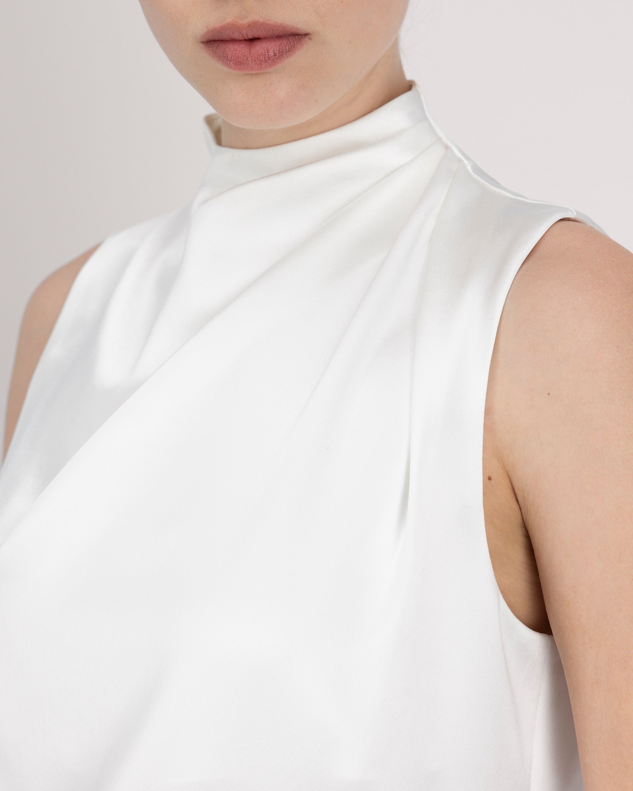 Asymmetrisch geschnittenes Top mit sanft drapierter Schulter für eine feminine, schwungvolle Silhouette. Der hochgeschlossene Stehkragen wird im Rückenteil durch einen feinen Schlitz und drei goldene Knöpfe mit filigranen Knopfschlingen akzentuiert. Ein edles Detail, das das puristische Design um eine subtile Note von Noblesse ergänzt.